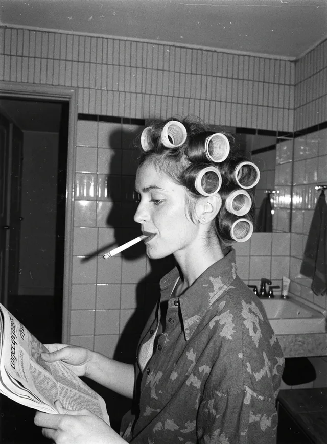 Femme aux bigoudis, une cigarette aux lèvres, lisant un journal dans une salle de bains