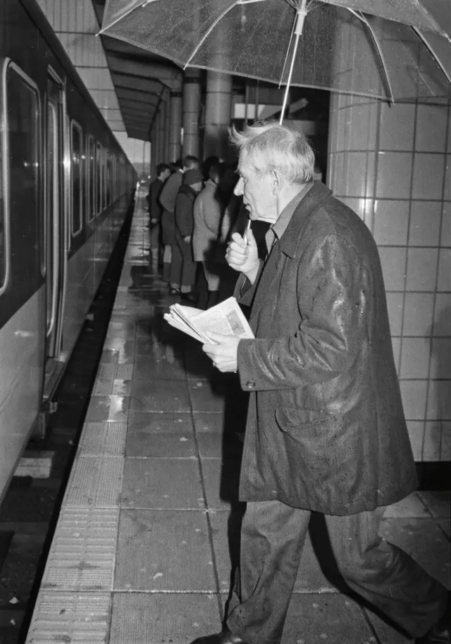 Homme au journal et au parapluie sur un quai de gare, en photographie noir et blanc